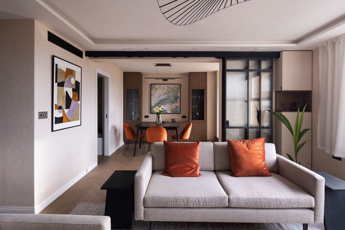 Living room with oatmeal sofa and terracotta cushions opening onto a dining area behind black steel-framed glazing – Boydell Court by Hone Studio. Photograph by Daniel Avery.