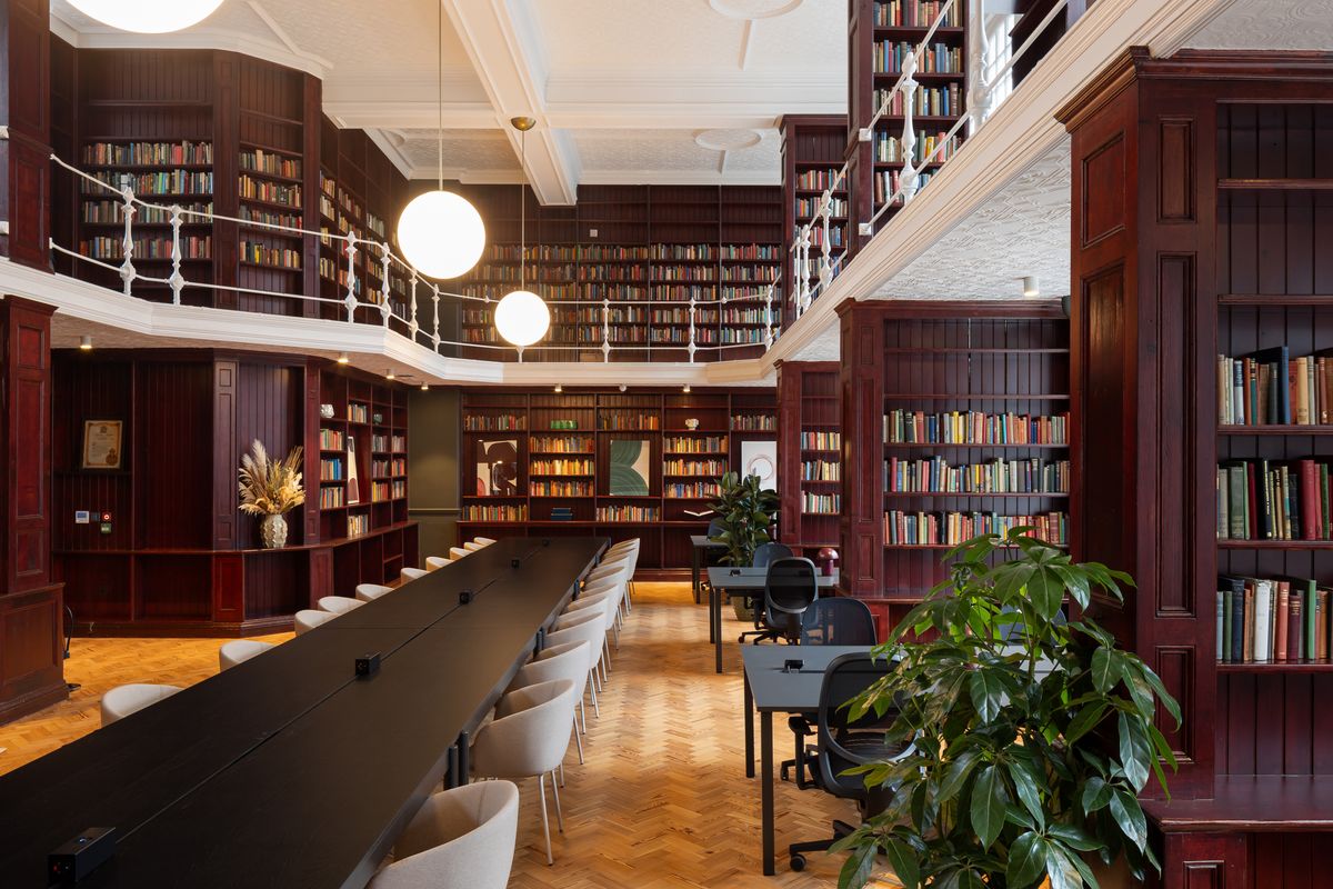 Double-height library workspace with burgundy bookcases, gallery balcony and long shared desk – Walworth Town Hall by Feix&Merlin. Photograph by Daniel Avery.
