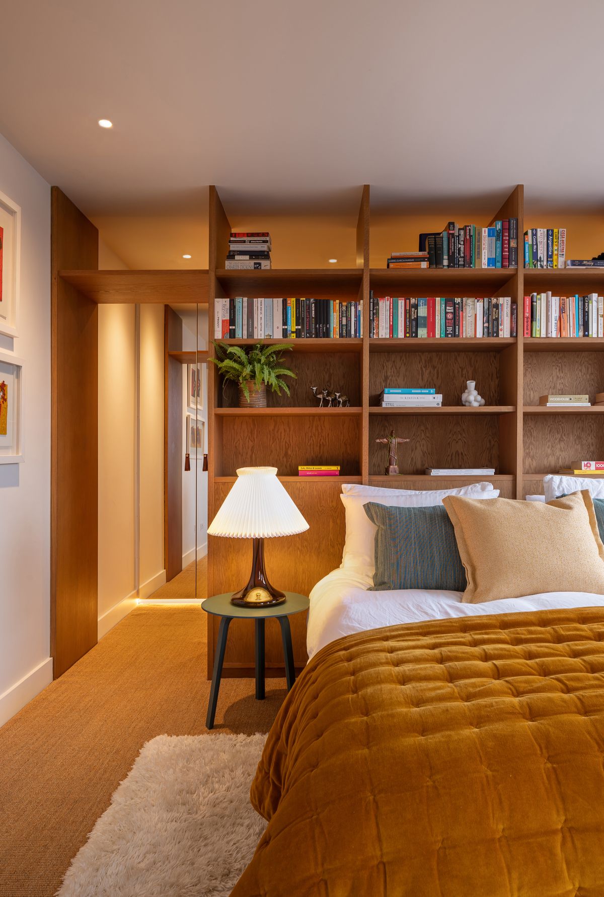Bedroom with built-in oak bookshelves, ochre velvet bedspread and table lamp – Overstrand Mansion by Studio Von B. Photograph by Daniel Avery.